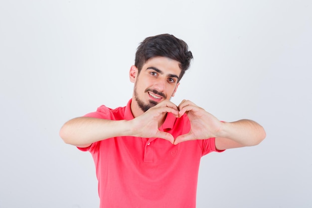 Young male showing heart gesture in pink t-shirt and looking confident. front view.