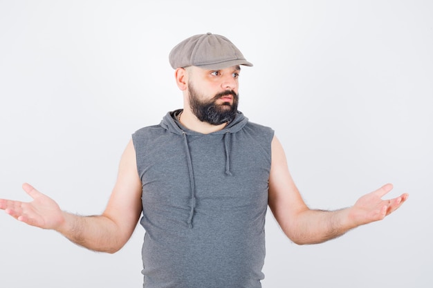 Young male showing helpless gesture in sleeveless hoodie, cap and looking desperate. front view.