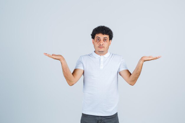 Young male showing helpless gesture in white t-shirt, pants and looking puzzled , front view.