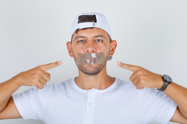 Free photo young male showing his taped mouth in white t-shirt, cap and looking calm, front view.