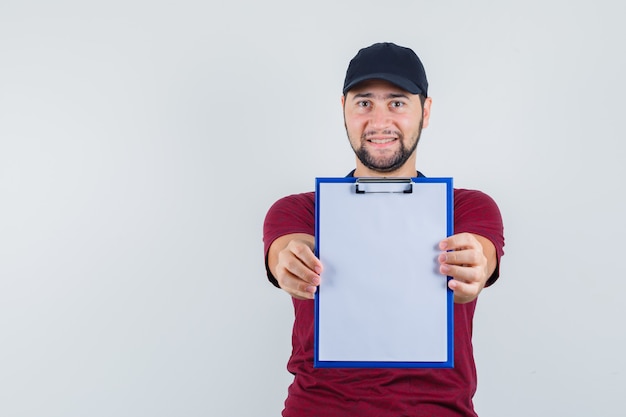 Free photo young male showing notebook in red t-shirt,black cap and looking glad , front view.