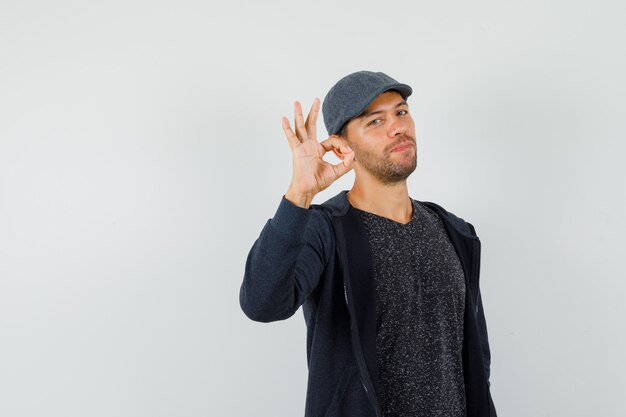 Young male showing ok gesture in t-shirt, jacket, cap and looking confident , front view.