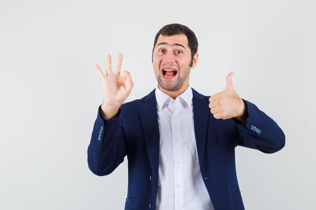 Young male showing ok gesture with thumb up in shirt and jacket and looking happy