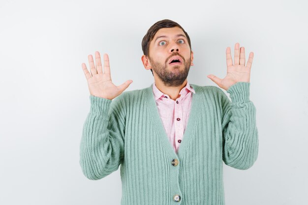 Young male showing palms in surrender gesture in shirt, cardigan and looking scared. front view.