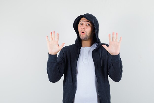 Young male showing palms in surrender gesture in t-shirt, jacket and looking scared. front view.