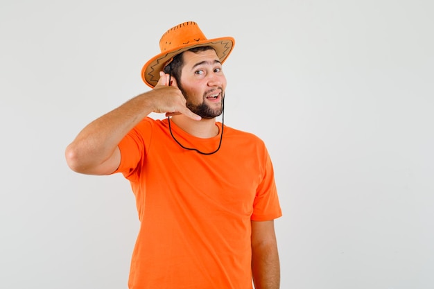 Молодой мужчина показывает телефонный жест в оранжевой футболке, шляпе и выглядит веселым, вид спереди.
