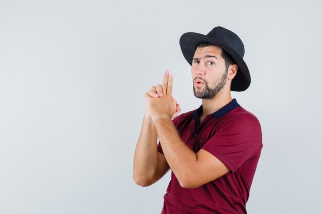 Young male showing pistol sign in t-shirt,hat and looking brave. front view.