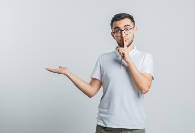 Young male showing silence gesture, spreading palm aside in white t-shirt and looking careful