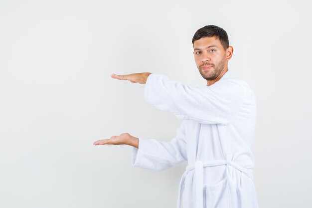Young male showing size sign and smiling in white bathrobe front view.