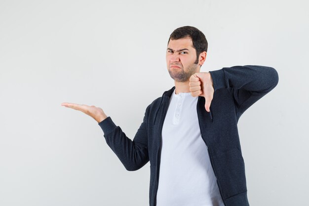 Young male showing something with thumb down in t-shirt, jacket and looking dissatisfied. front view.