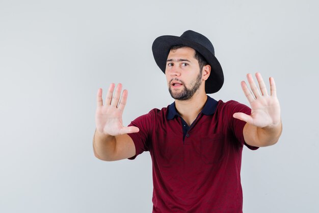 Young male showing stop gesture in t-shirt,hat and looking excited. front view.