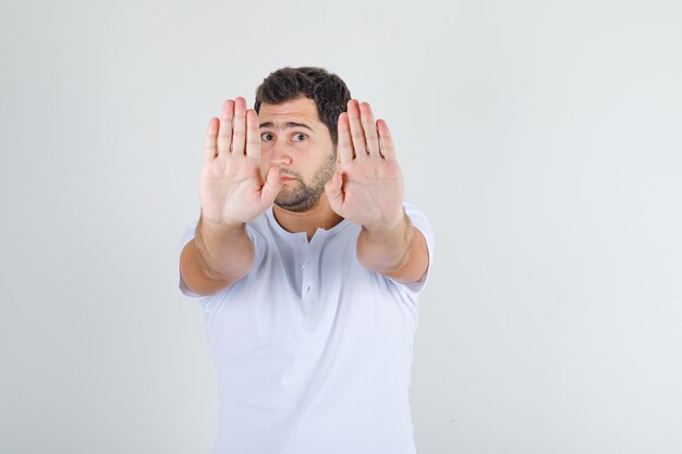 Young male showing stop gesture in white t-shirt and looking disgusted