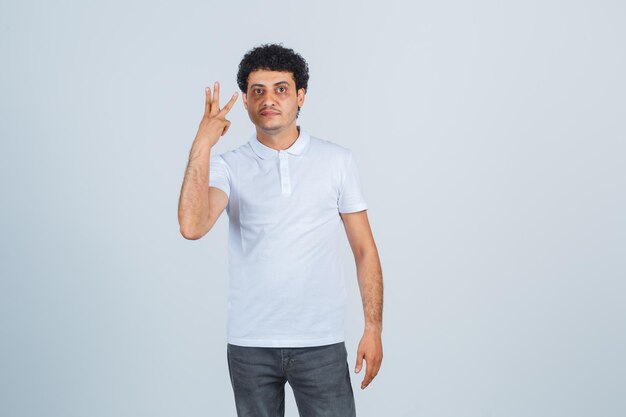 Молодой мужчина показывает три пальца в белой футболке, брюках и выглядит уверенно, вид спереди.