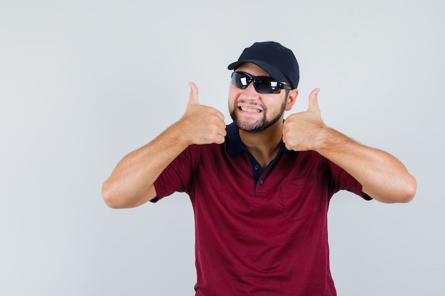 Young male showing thumb up in t-shirt,sunglasses and looking glad , front view.