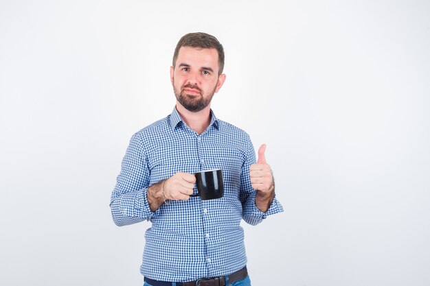 Young male showing thumb up while holding cup in shirt, jeans and looking delighted , front view.