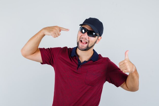 Young male showing thumb up while pointing at his glasses in t-shirt,black glasses and looking satisfied , front view.