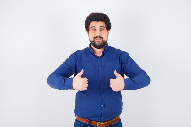 Young male showing thumb ups in shirt, jeans and looking happy , front view.