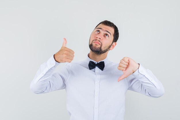 Young male showing thumbs down and up in white shirt and looking confused , front view.