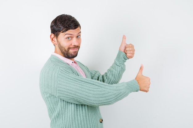 Young male showing thumbs up in shirt, cardigan and looking confident. front view.