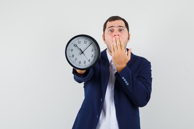 Young male showing wall clock in shirt and jacket and looking scared