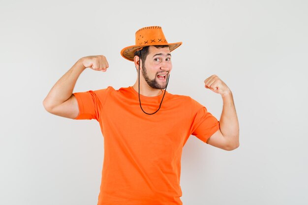 Young male showing winner gesture in orange t-shirt, hat and looking joyful. front view.
