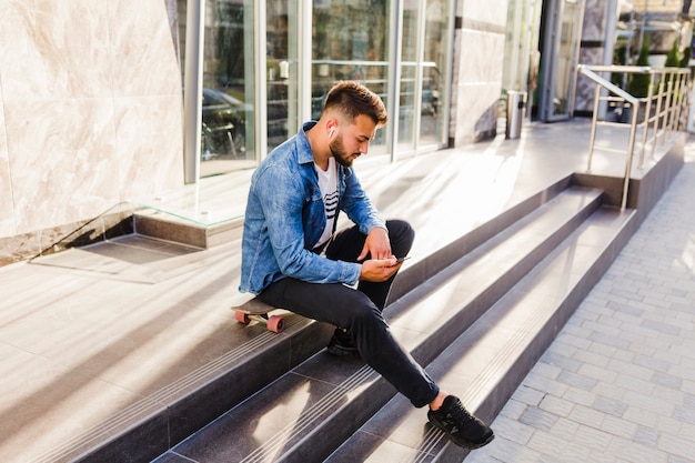 Young male skateboarder sitting on skateboard using cellphone