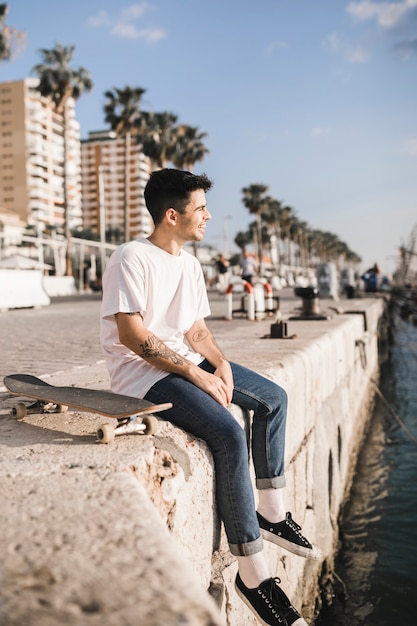 Young male skateboarder with a skateboard sitting on retaining wall