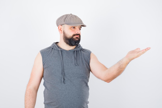 Free photo young male in sleeveless hoodie, cap raising hand in questioning pose and looking confident , front view.