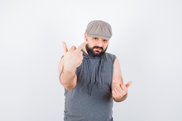 Free photo young male in sleeveless hoodie, cap showing middle fingers and looking confident , front view.