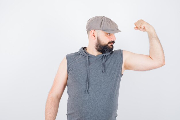 Young male in sleeveless hoodie, cap showing muscles of arm and looking confident , front view.