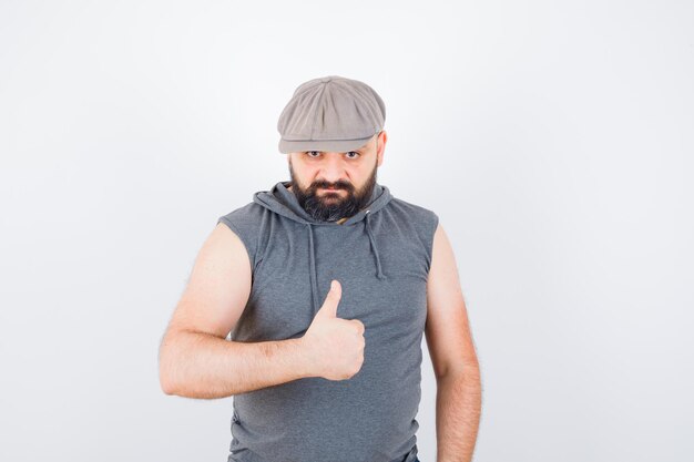 Young male in sleeveless hoodie, cap showing thumb up and looking confident , front view.