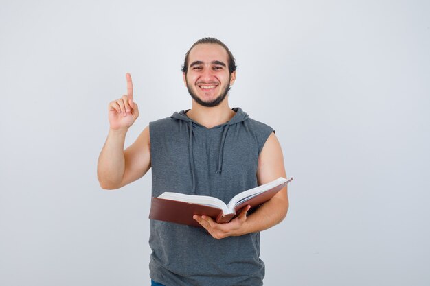 Young male in sleeveless hoodie holding book while pointing up and looking happy , front view.