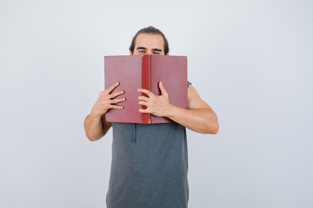Young male in sleeveless hoodie holding opened book on face and looking sleepy , front view.