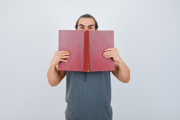 Young male in sleeveless hoodie holding opened book on mouth and looking serious , front view.