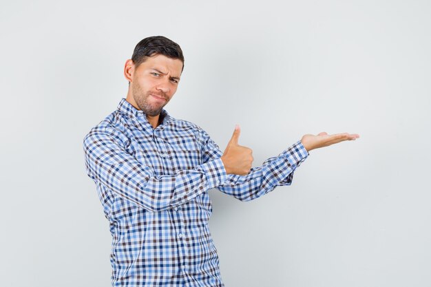 Young male spreading palm aside, showing thumb up in checked shirt and looking confident