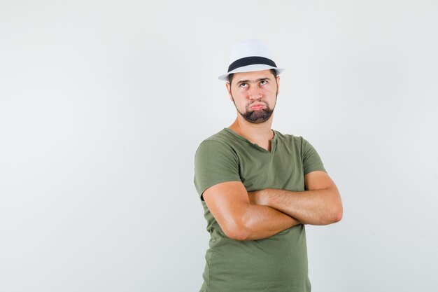 Young male standing with crossed arms in green t-shirt and hat and looking irritated