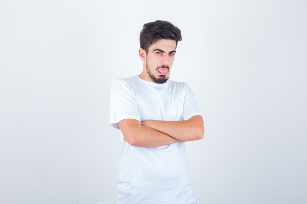 Young male standing with crossed arms while sticking out tongue in t-shirt and looking funny , front view.