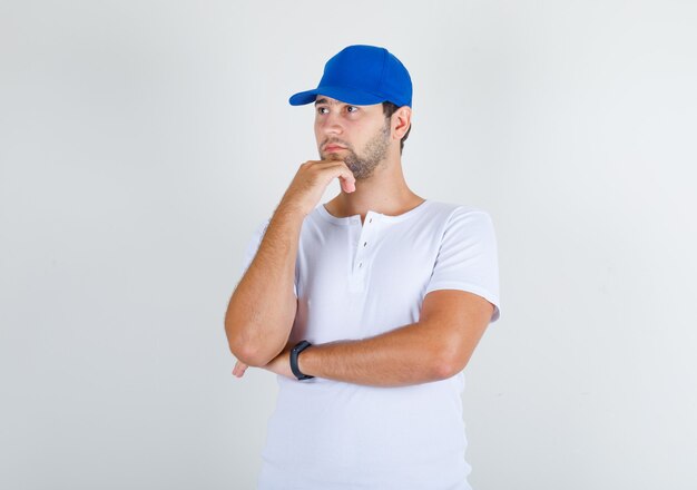 Молодой мужчина стоит с рукой на подбородке в белой футболке и синей кепке