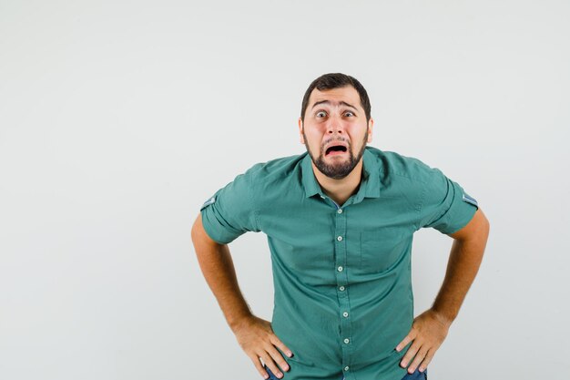 Young male standing with hands on waist in green shirt and looking distressed. front view.