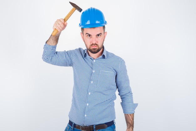 Giovane maschio che colpisce la testa con un martello in camicia, jeans, casco e sembra stupido, vista frontale.