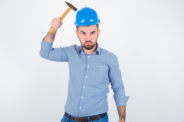 Young male striking his head with a hammer in shirt, jeans, helmet and looking stupid , front view.