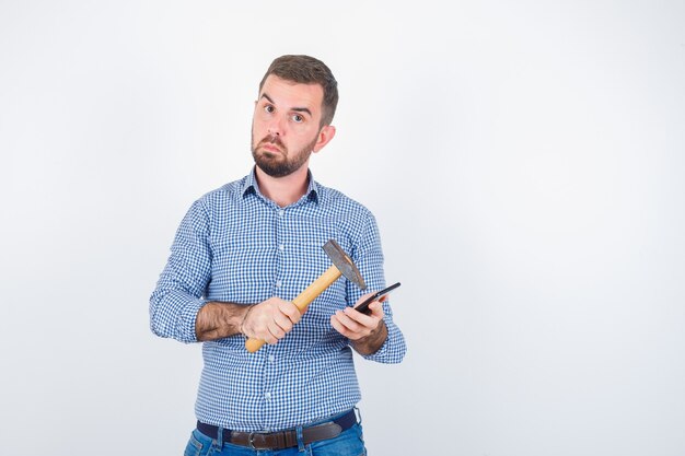 Young male striking mobile phone with a hammer in shirt, jeans and looking hesitant , front view.