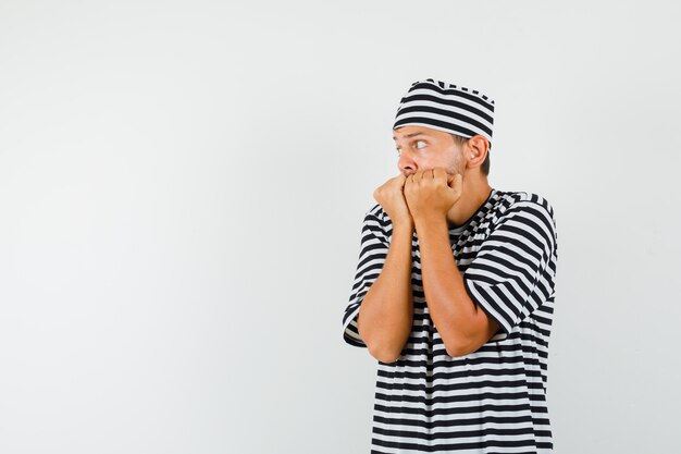 Young male in striped t-shirt hat biting fists emotionally and looking terrified  