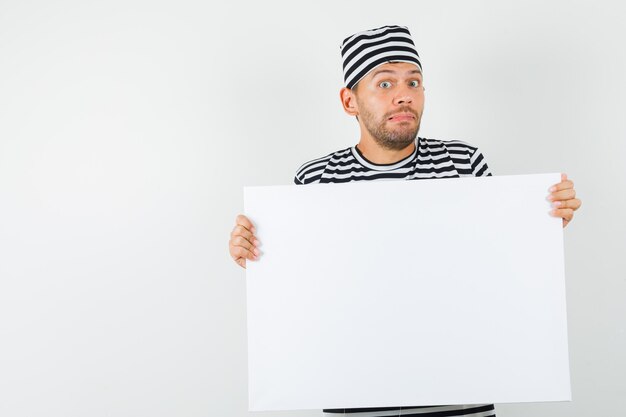 Young male in striped t-shirt hat holding blank canvas and looking confused  