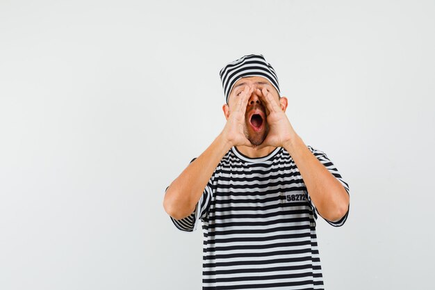 Young male in striped t-shirt hat telling secret with hands near open mouth  