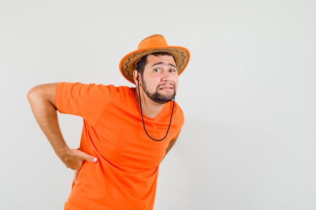 오렌지색 티셔츠, 모자를 쓰고 요통을 앓고 있는 젊은 남성이 피곤해 보입니다. 전면보기.