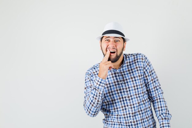 Young male suffering from painful toothache in checked shirt, hat and looking uncomfortable. front view.