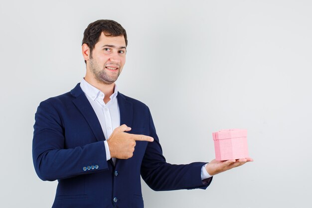 Young male in suit pointing finger at gift box and looking cheery , front view.