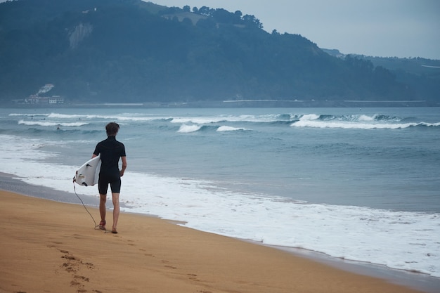 잠수복에 젊은 남성 서퍼가 파도를보고 자신의 흰색 서핑 보드와 함께 해변을 따라 산책