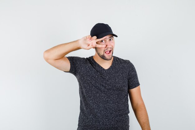 Young male in t-shirt and cap holding v-sign on eye